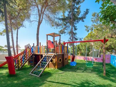 Bunter Spielplatz mit Rutsche, Klettergerüst und zwei Schaukeln unter Bäumen auf grünem Rasen.