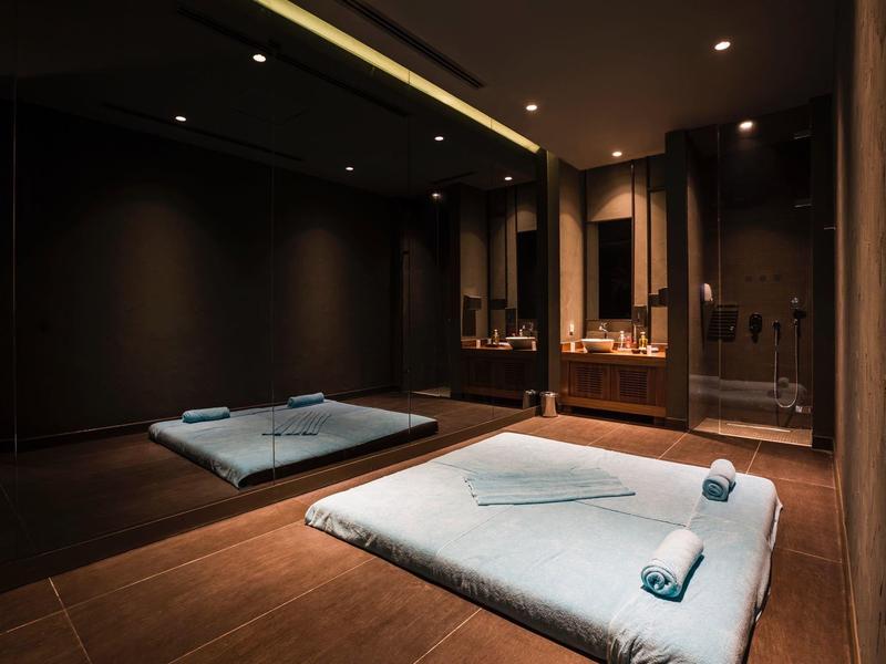 Pohodlná spa místnost s dvěma podlahovými podložkami, tmavými stěnami, jemným osvětlením a srolovanými ručníky.