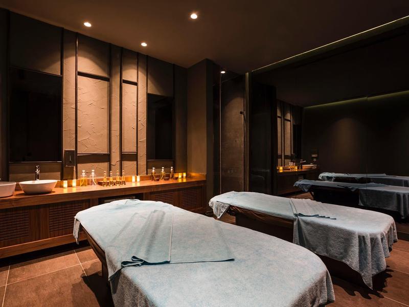 Slabě osvětlená spa místnost se dvěma masážními stoly přikrytými bílými plachtami a nástěnnou skříní se svíčkami.