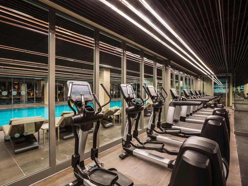 Moderní posilovna s běžeckými pásy a eliptickými trenažéry vedle velkého okna odrážejícího bazén venku.