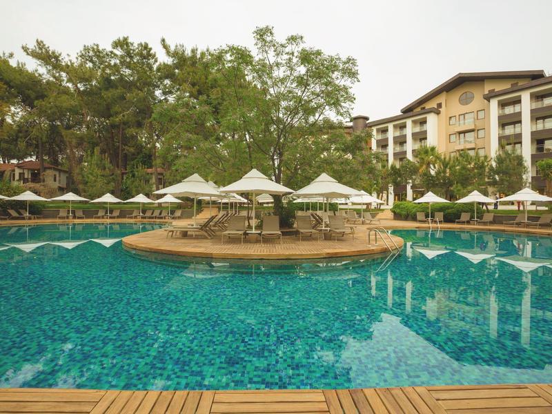 Velký bazén s modrou vodou, slunečníky a hotelem v pozadí.