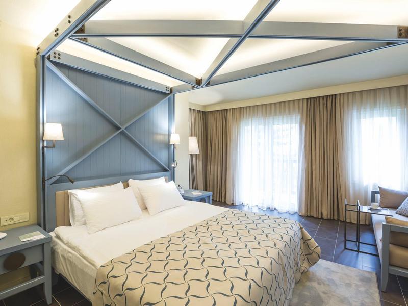 Jasný hotelový pokoj s velkou postelí, geometrickou čelní stěnou, závěsy a útulným sezením.