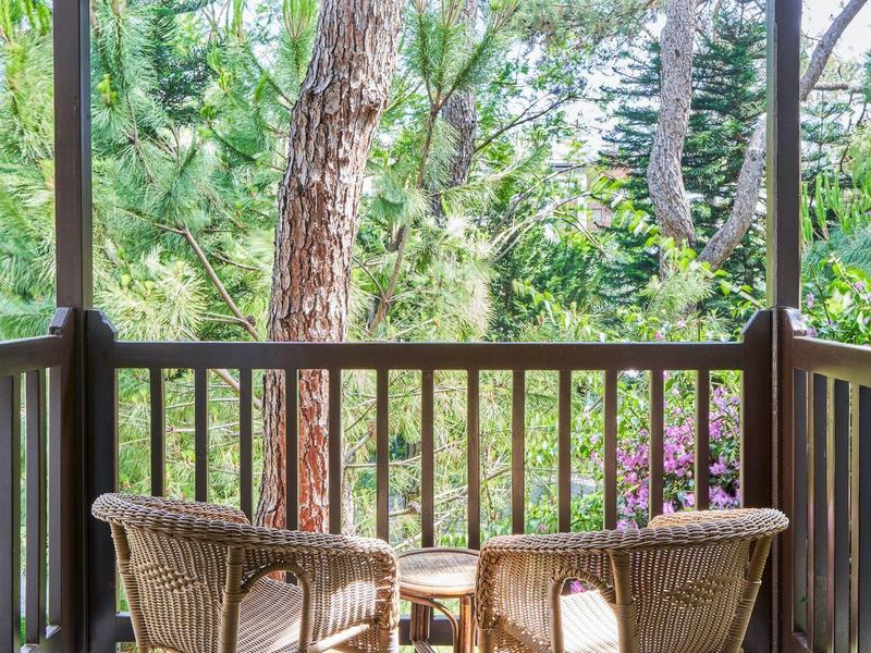 Dva proutěné židle a malý stolek na dřevěném balkóně s výhledem na stromy.