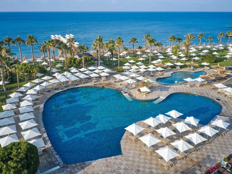 Bazénová oblast resortu s lehátky, slunečníky, palmami a výhledem na oceán za jasné oblohy.