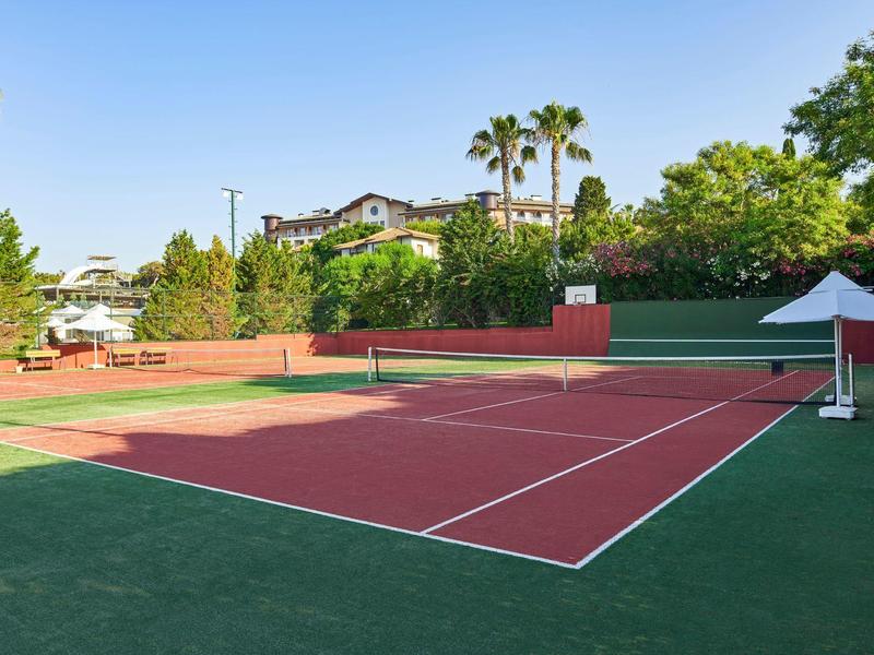 Venkovní tenisový kurt s červenou a zelenou plochou, obklopený stromy a slunečníky pod modrou oblohou.