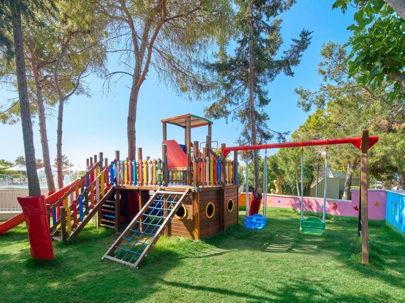 Barevné dřevěné dětské hřiště ve tvaru lodi na zelené trávě pod vysokými stromy.