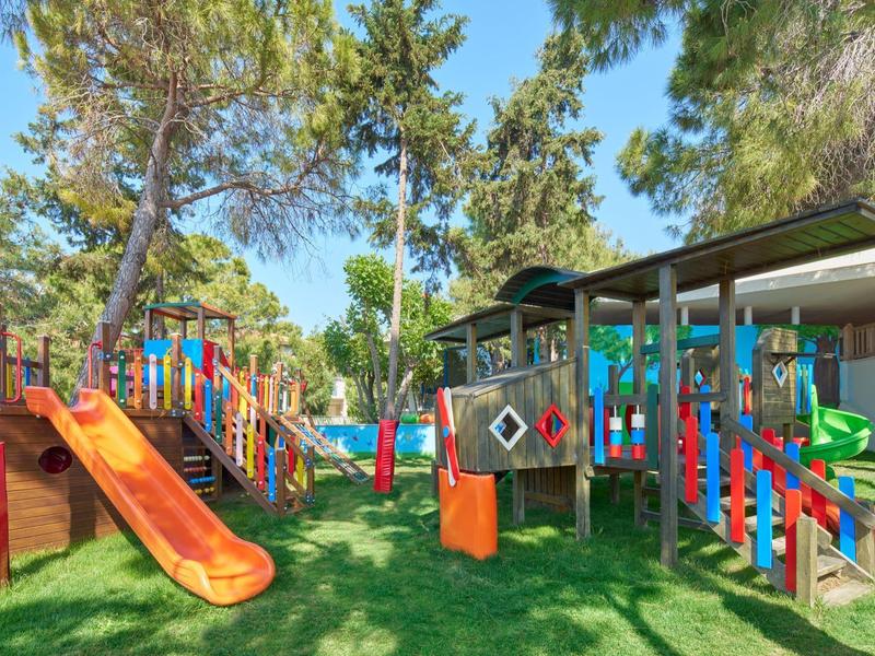 Venkovní hřiště s barevnými skluzavkami a prolézačkami na zelené trávě pod stromy.