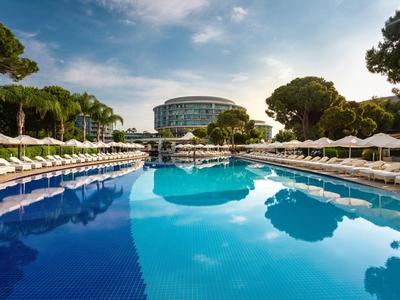 Großer blauer Pool mit Liegestühlen und Sonnenschirmen, umgeben von Bäumen und einem Hotelgebäude.