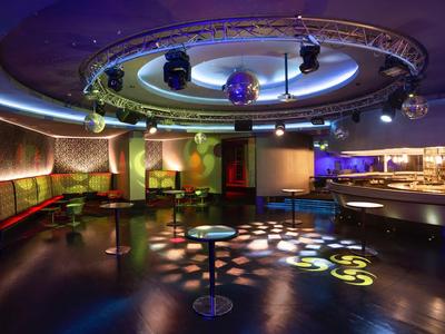 Moderner Nachtclub mit runden Tischen, Bar, Disco-Kugel und farbigen Lichtreflexen auf dem Boden.
