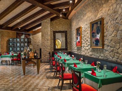 Rustikales Restaurant mit Steinwänden, Holzbalkendecke, grünen Tischdecken und roten Stühlen.