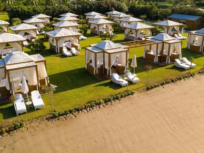 Mehrere weiße Strandpavillons mit Liegestühlen stehen auf grüner Wiese am Sandstrand.