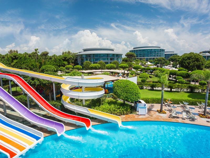 Toboganes de colores desembocan en una piscina frente a un hotel moderno con jardines verdes.