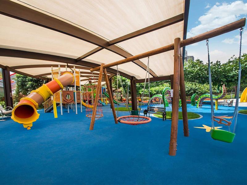 Parque infantil cubierto con tobogán, columpios y suelo blando en un jardín de hotel.