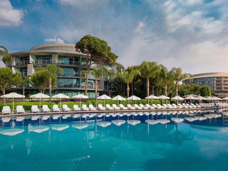 Hotel de lujo con piscina, tumbonas y palmeras bajo un cielo azul.