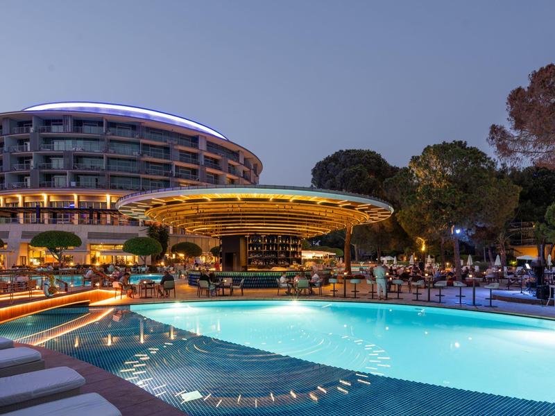 Piscina de hotel iluminada y edificio redondo al anochecer con invitados y árboles