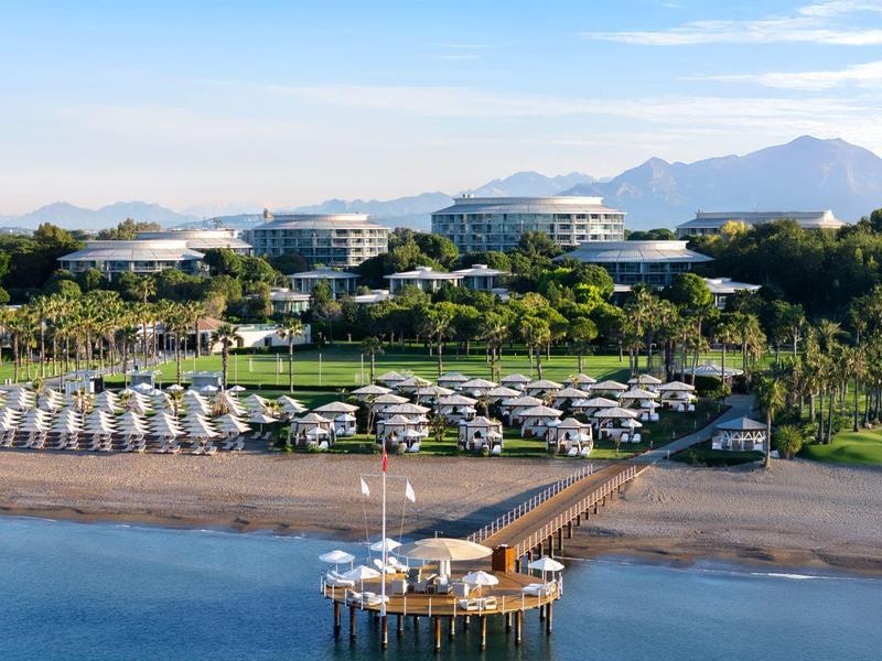 Hotel de playa con tumbonas, sombrillas y muelle sobre un mar tranquilo bajo cielo azul.