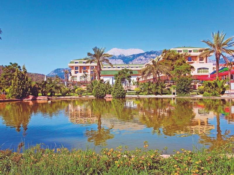 Resortgebouwen met palmbomen bij een rustig meer met reflectie van blauwe lucht en bergen.