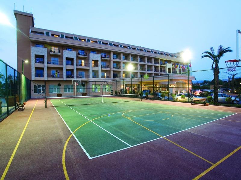 Tennisbaan naast een modern hotelgebouw in de schemering met verlichting en palmbomen in de buurt.