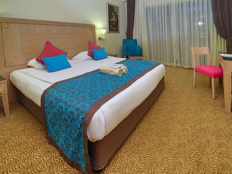 Hotelkamer met groot bed, kleurrijke kussens, bureau, stoel en raam met gordijnen.