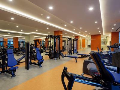 Moderne Fitnesshalle mit verschiedenen Trainingsgeräten, blauen Sitzen und hellem Deckenlicht.