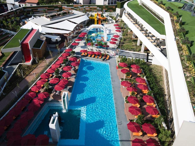Vista aerea di una piscina d'hotel con ombrelloni rossi e lettini accanto a un edificio moderno.