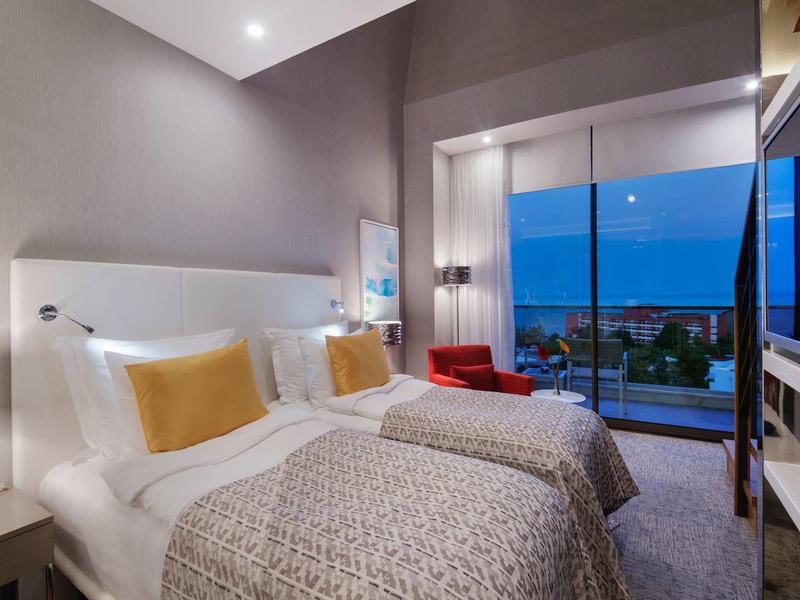 Camera d'albergo moderna con letto matrimoniale, balcone e vista mare al crepuscolo.