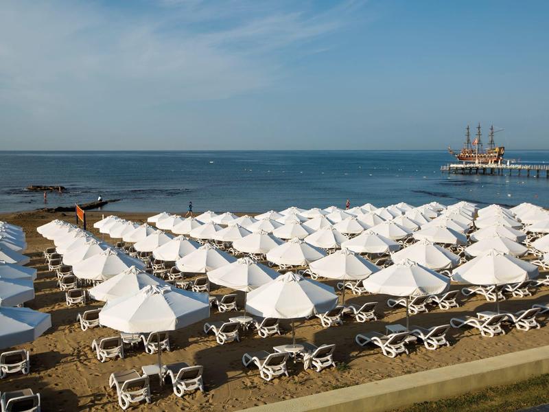 Spiaggia con file di ombrelloni bianchi e lettini vicino al mare, veliero in lontananza.