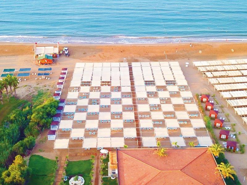 Vista aerea di una grande scacchiera sulla spiaggia accanto a un edificio con tetto rosso e il mare blu sullo sfondo.