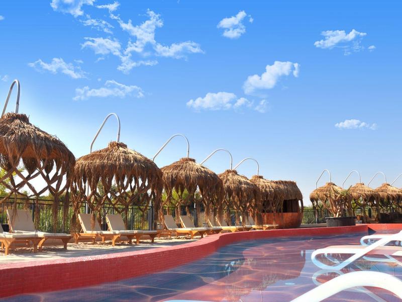 Piscina rossa con sedie a sdraio bianche e tetti di paglia tradizionali sotto un cielo blu.