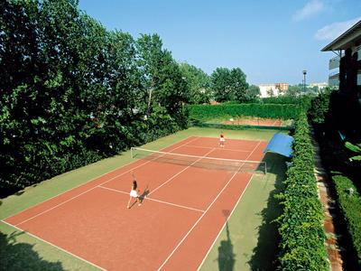 Court de tennis extérieur en terre battue rouge entouré d'arbres et de bâtiments sous un ciel bleu.