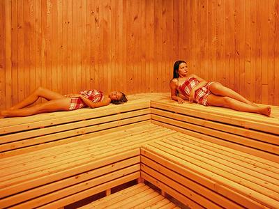 Twee vrouwen ontspannen op houten banken in een lichte sauna met houten panelen.