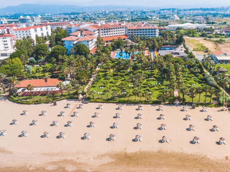Luchtfoto van een strand met ligstoelen, op de achtergrond een hotel en groene tuinen.