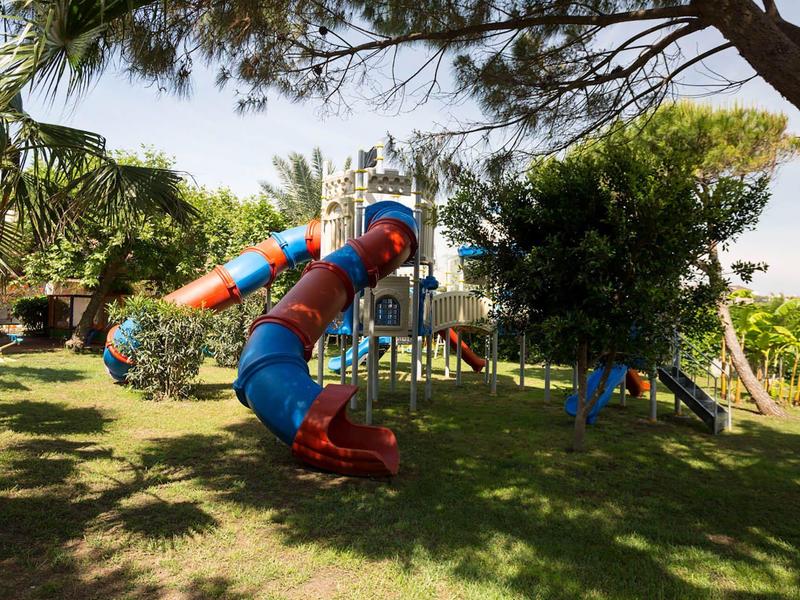 Speeltuin met kleurrijke glijbanen en klimrekken op gras onder bomen.