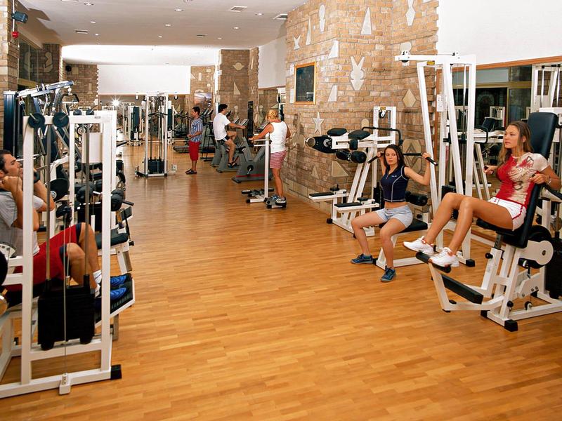 Sportschool met houten vloer en meerdere fitnessapparaten, mensen aan het sporten.