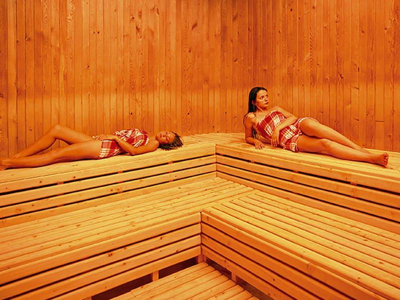 Twee vrouwen ontspannen op houten banken in een lichte sauna met houten panelen.