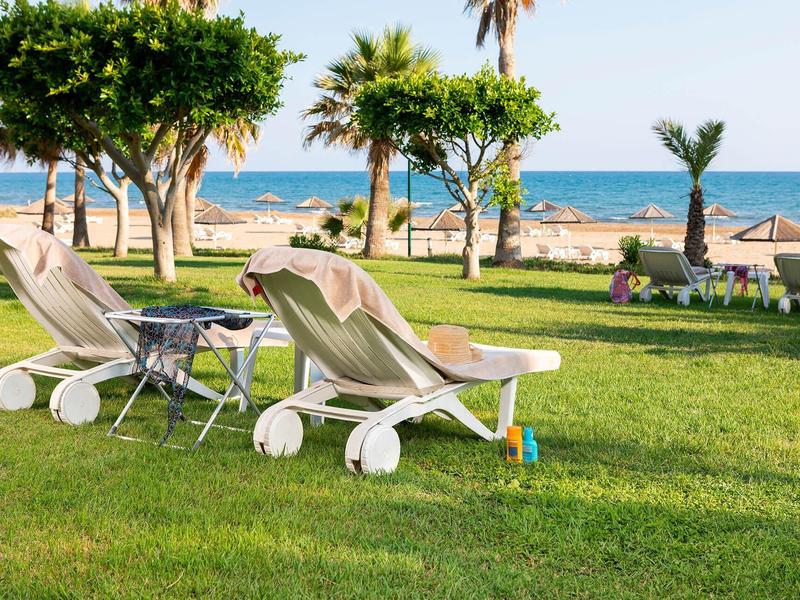 Twee ligstoelen met tafels op een groen gazon aan zee met palmbomen op de achtergrond.