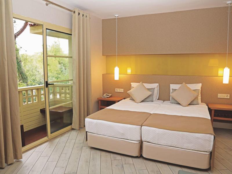 Bright hotel room with two beds and access to a balcony overlooking greenery.