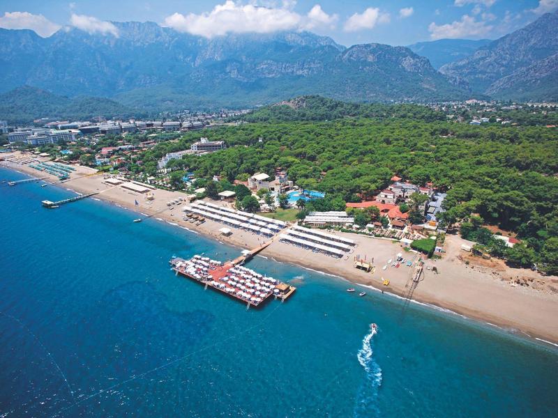 Aerial view of a beach with blue sea, piers, and a wooded hotel area in the background.