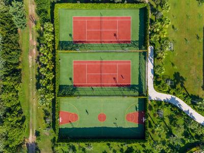 Trzy ogrodzone boiska sportowe z kortami tenisowymi i koszykówką otoczone zielonym krajobrazem.