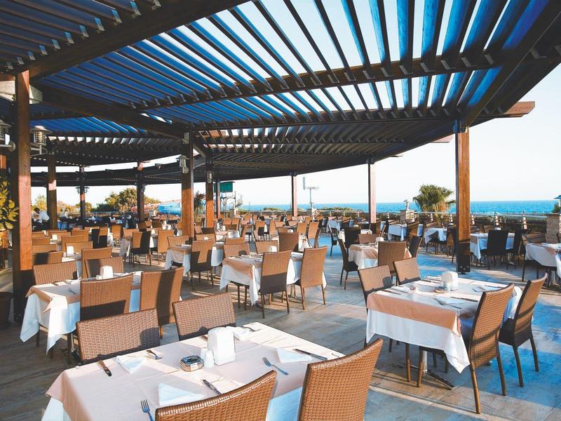 Duży taras z nakrytymi stołami i krzesłami z widokiem na morze i daszkiem przeciwsłonecznym.