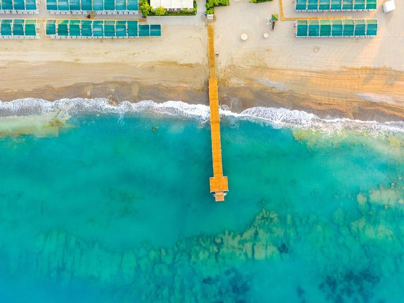 Widok z powietrza długiego pomostu na piaszczystej plaży z leżakami i turkusową wodą.