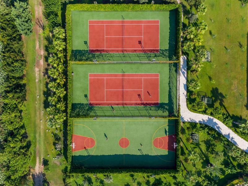 Trzy ogrodzone boiska sportowe z kortami tenisowymi i koszykówką otoczone zielonym krajobrazem.