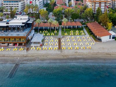 Strand mit gelben Liegestühlen, Gebäuden und Bäumen im Hintergrund, klares blaues Meer im Vordergrund.