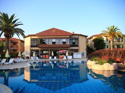 Großer blauer Pool vor zweistöckigem Gebäude mit roten Dächern, Palmen und Sonnenliegen.