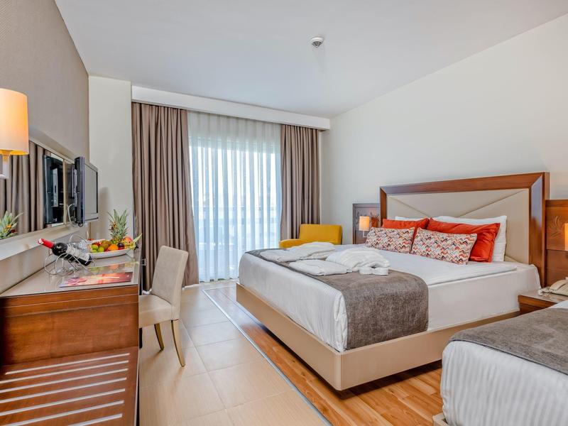 Bright hotel room with two beds, desk, chair, and balcony with curtains.