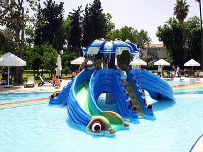 Kolorowy plac zabaw z zjeżdżalniami wodnymi w płytkim basenie z parasolami i drzewami w tle.