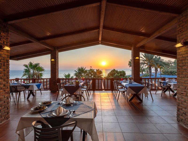 Zadaszony taras z nakrytymi stołami i widokiem na zachód słońca oraz palmy nad morzem.