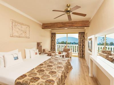 Hell eingerichtetes Hotelzimmer mit zwei Betten, Ventilator an der Decke und Balkon mit Blick auf Meer.