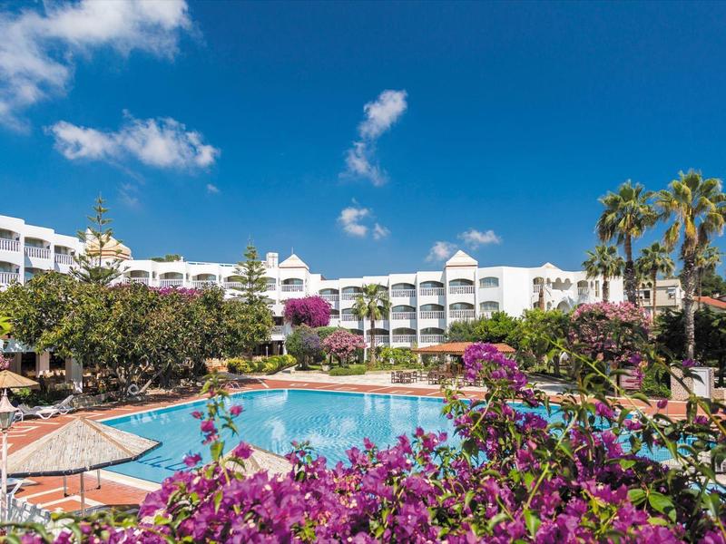 Großer Pool mit klar blauem Wasser, umgeben von Palmen und weißem Hotelgebäude unter blauem Himmel.