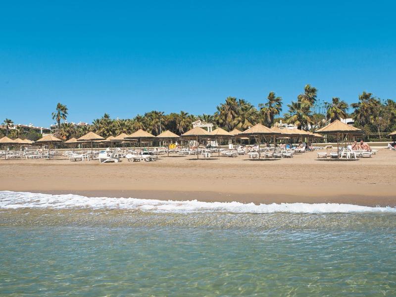 Strand mit klarem Wasser, Sand, Liegestühlen und Sonnenschirmen unter blauem Himmel.